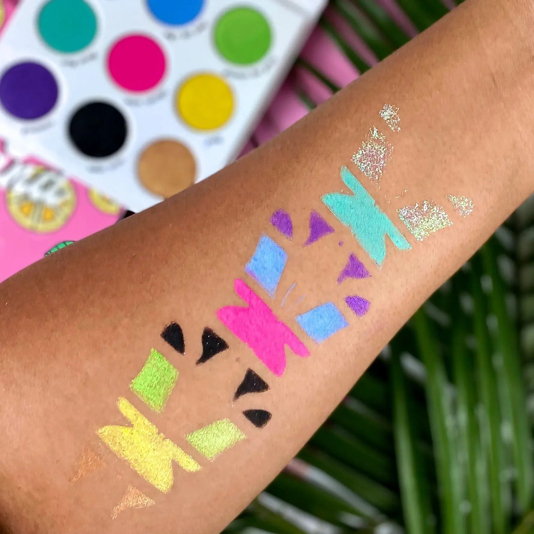 Colorful glittery designs on a person's arm with a makeup palette in the background.
