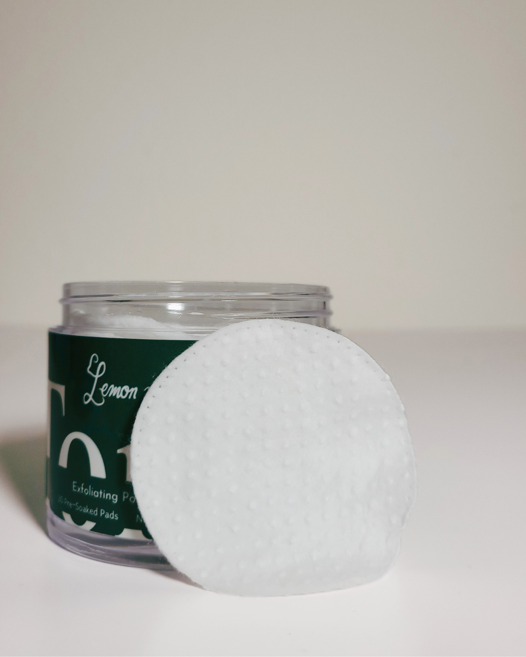 A clear jar of lemon-colored facial cleansing pads with a dark green label, next to a round white textured cotton pad on a plain light background.