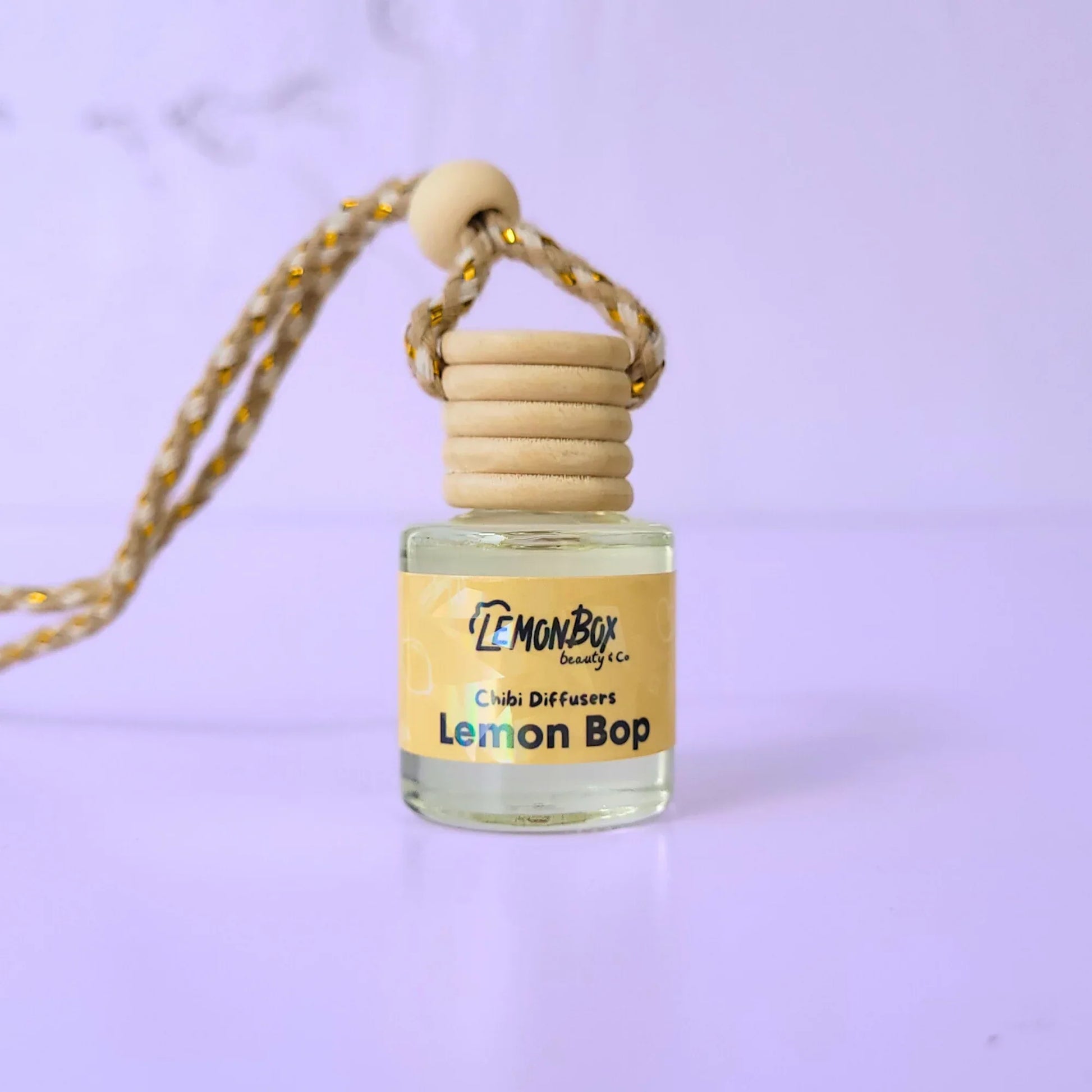 A small glass Chibi Diffuser bottle labeled "Lemon Bop" with a wooden screw-on cap and a tan braided hanging cord, set against a soft purple background.