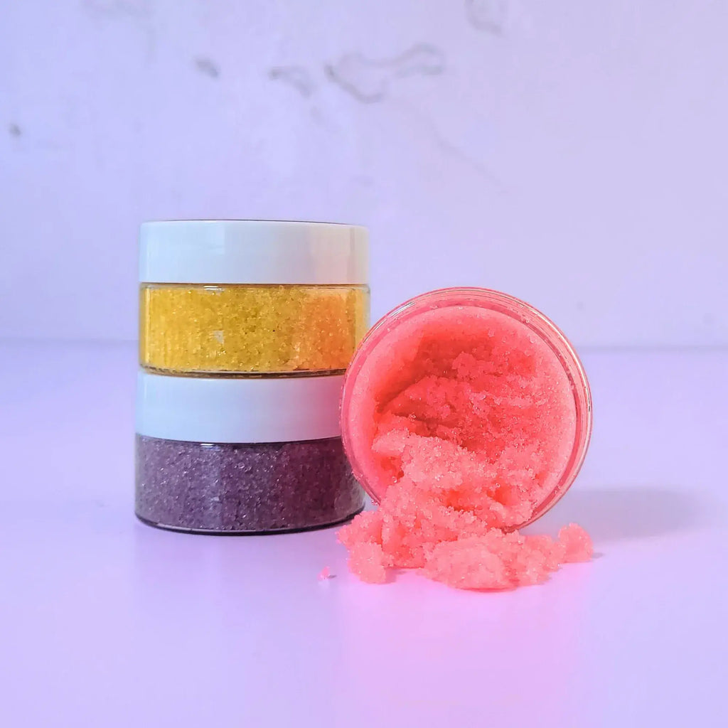 Close-up of Lemon Box Beauty pink Strawberry Coconut lip scrub texture on a spoon, with a stack of yellow and purple sugar scrubs in the background.