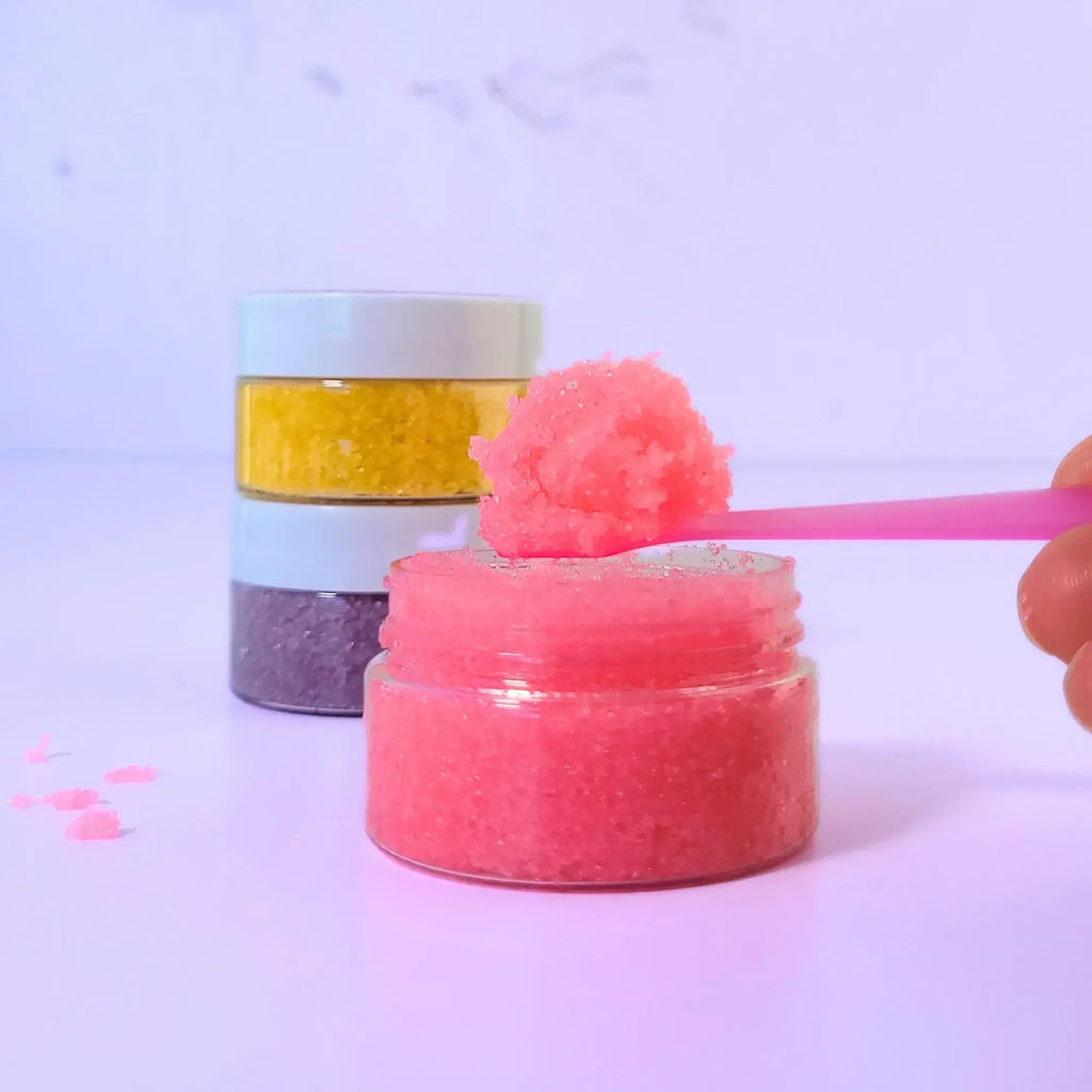 Close-up of Lemon Box Beauty pink Strawberry Coconut lip scrub texture on a spoon, with a stack of yellow and purple sugar scrubs in the background.