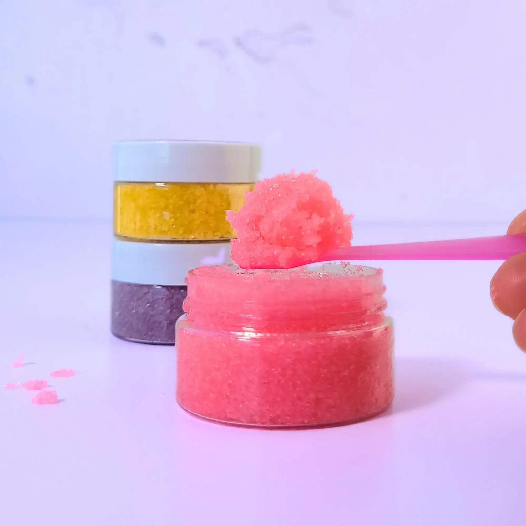 Close-up of Lemon Box Beauty pink Strawberry Coconut lip scrub texture on a spoon, with a stack of yellow and purple sugar scrubs in the background.