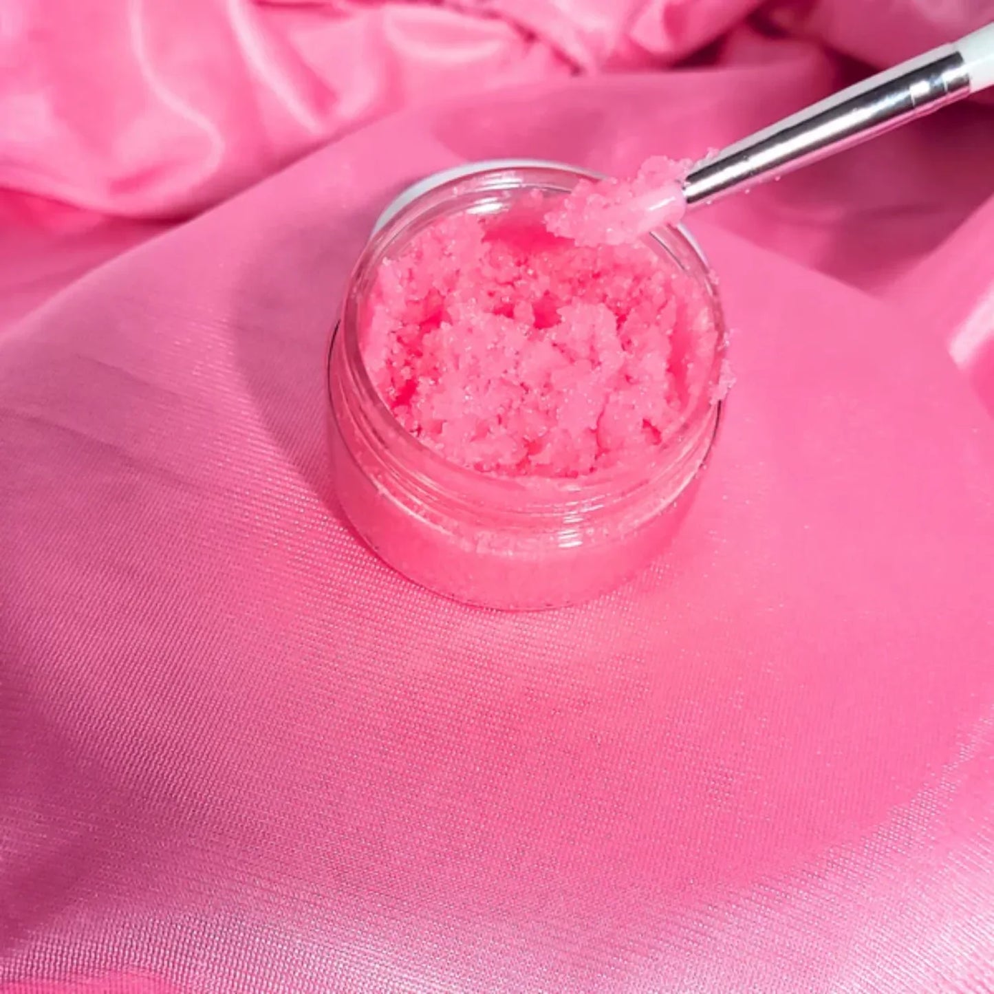 Pink sugar scrub in a jar with a spoon on pink fabric