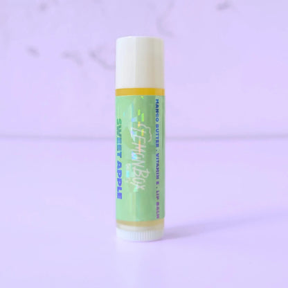 Lemon Box Beauty Sweet Apple Mango Butter and Vitamin E Lip Balm—a white and yellow tube with a holographic green label featuring blue and teal apple typography on a lavender background.