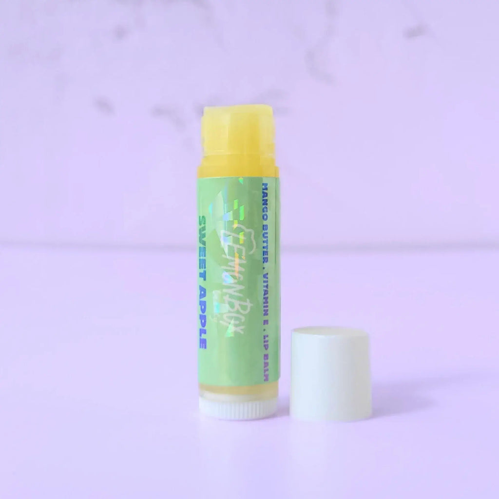 Lemon Box Beauty Sweet Apple Mango Butter and Vitamin E Lip Balm—a white and yellow tube with a holographic green label featuring blue and teal apple typography on a lavender background.