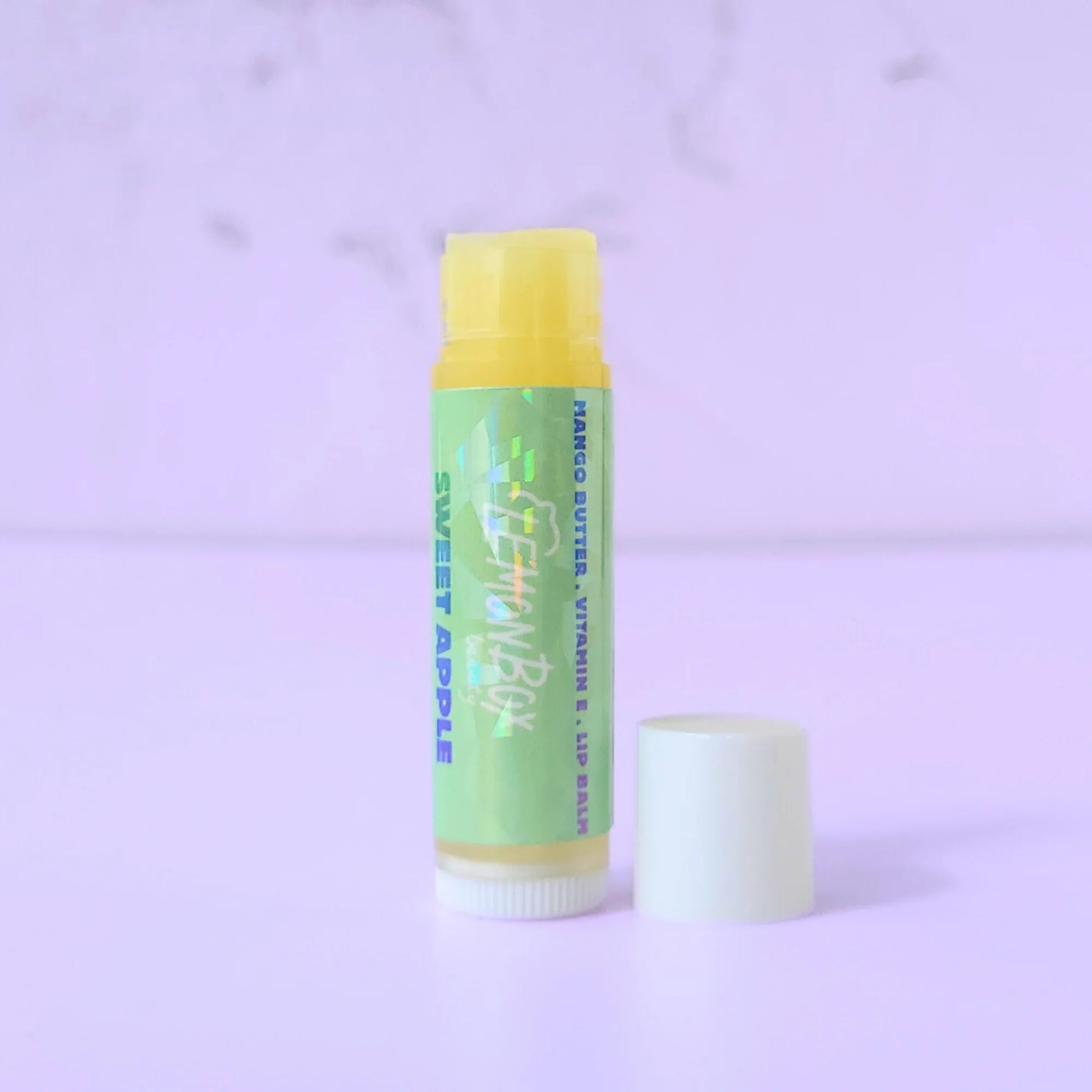 Lemon Box Beauty Sweet Apple Mango Butter and Vitamin E Lip Balm—a white and yellow tube with a holographic green label featuring blue and teal apple typography on a lavender background.