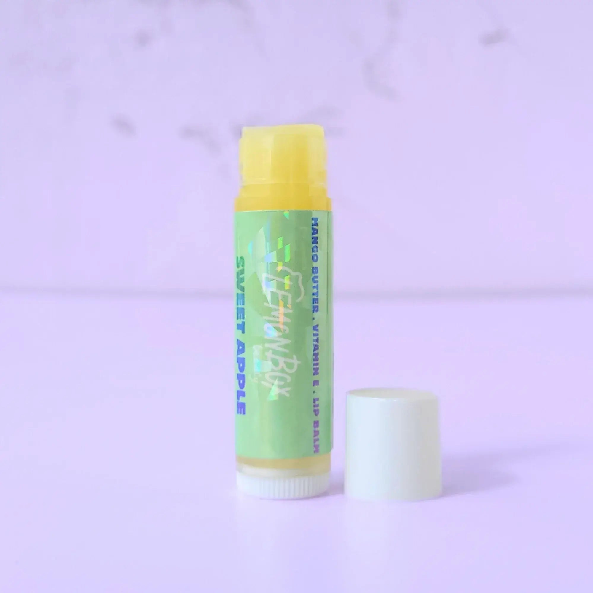 Lemon Box Beauty Sweet Apple Mango Butter and Vitamin E Lip Balm—a white and yellow tube with a holographic green label featuring blue and teal apple typography on a lavender background.