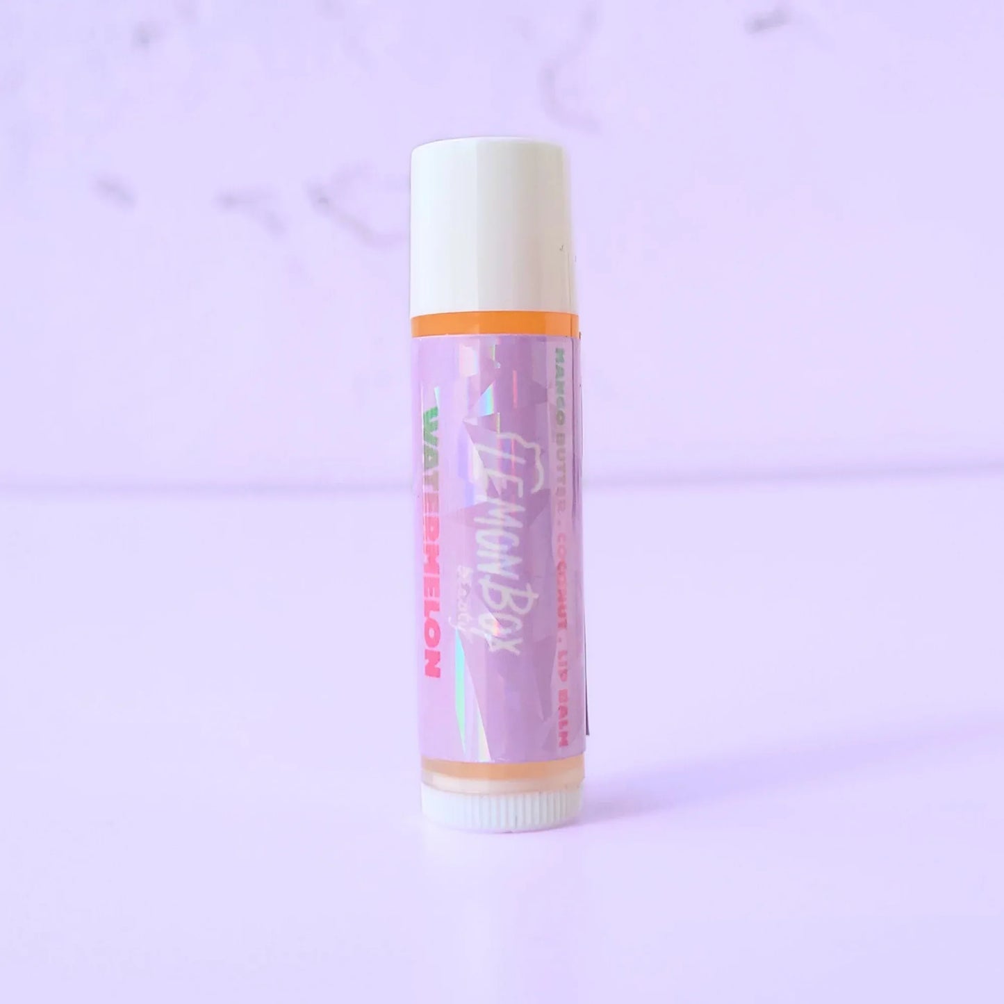 Lemon Box Beauty Watermelon Mango Butter and Vitamin E Lip Balm—a white and orange tube with a holographic purple label featuring green and pink watermelon typography on a lavender background.