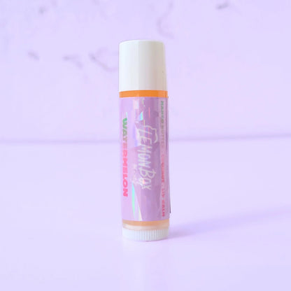 Lemon Box Beauty Watermelon Mango Butter and Vitamin E Lip Balm—a white and orange tube with a holographic purple label featuring green and pink watermelon typography on a lavender background.