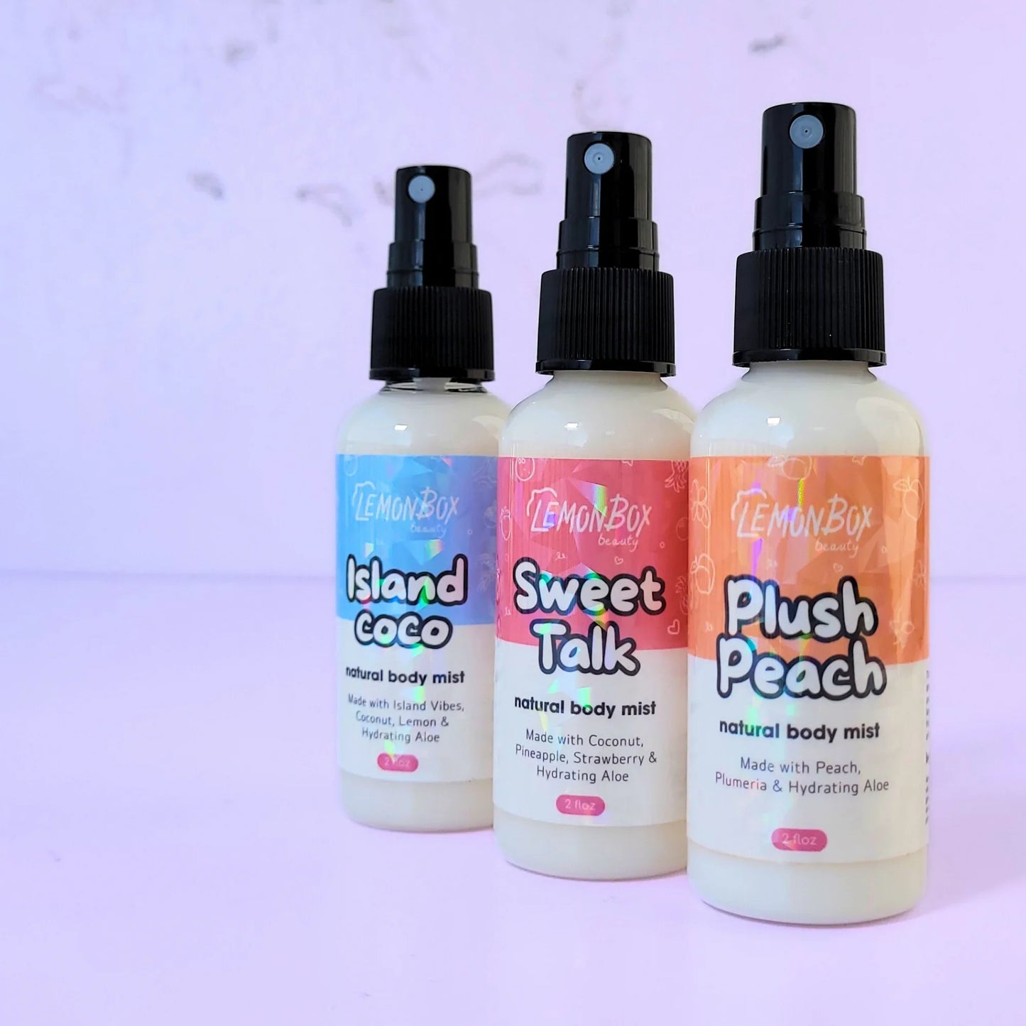 A trio of Lemon Box Beauty Natural Body Mists—Island Coco, Sweet Talk, and Plush Peach—2oz clear spray bottles with holographic labels arranged on a lavender background