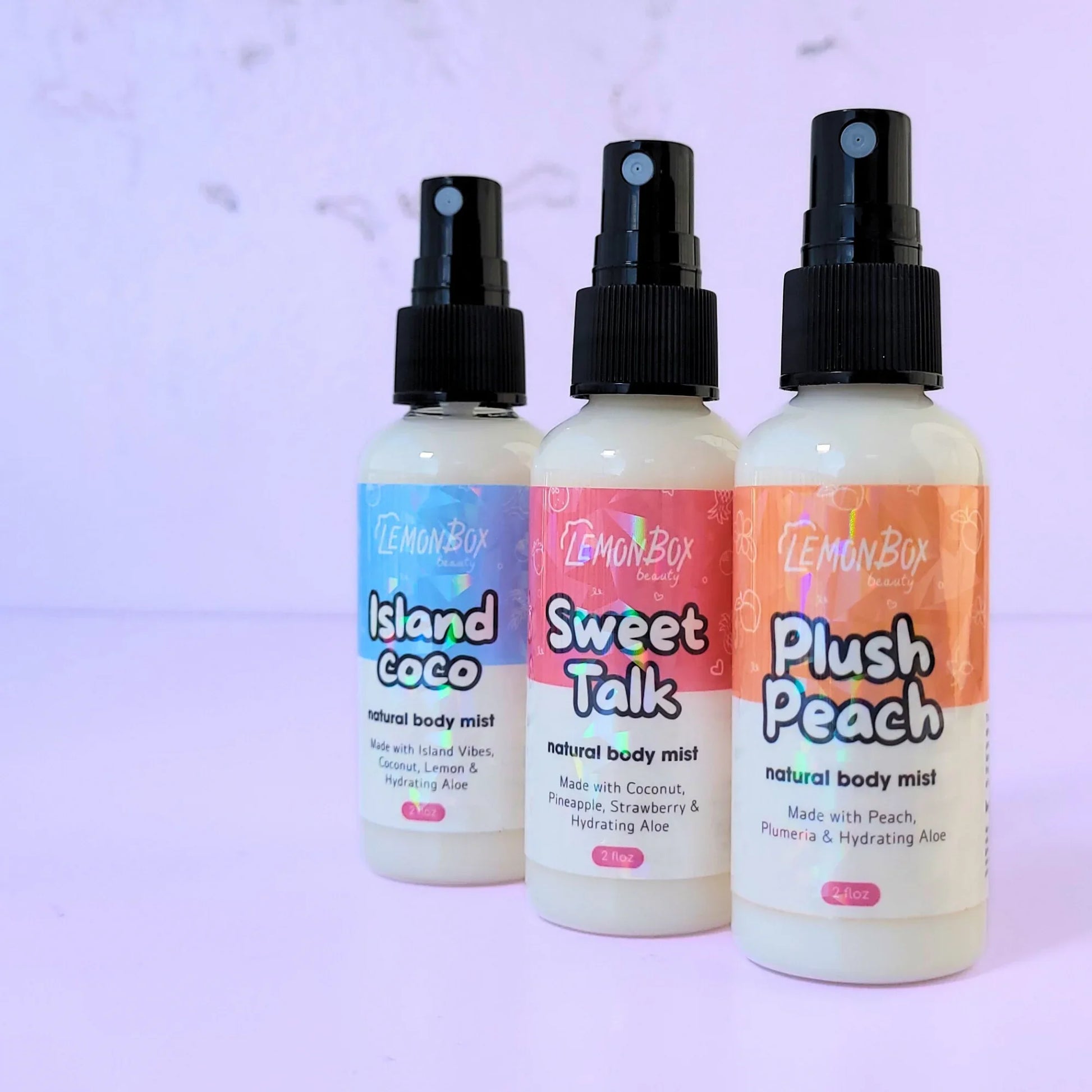 A trio of Lemon Box Beauty Natural Body Mists—Island Coco, Sweet Talk, and Plush Peach—2oz clear spray bottles with holographic labels arranged on a lavender background