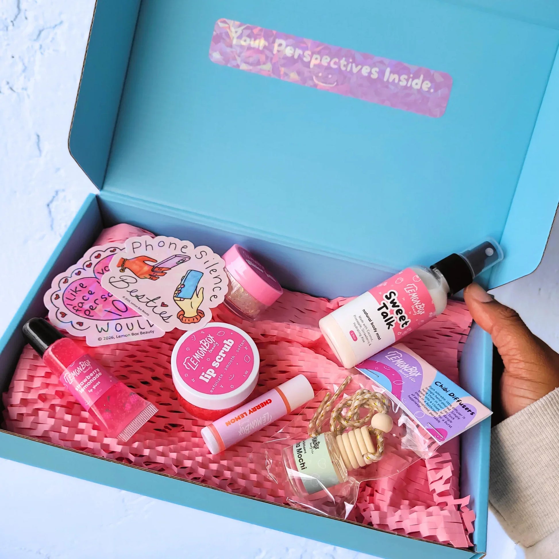 Open blue box with pink and white products including lip balm, lip gloss, lip scrub, oil diffuser, and other small items on a light blue background.